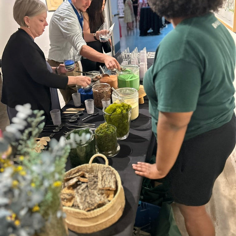 People preparing sand art terrariums at a table with various items including moss, sand, tools, and containers.